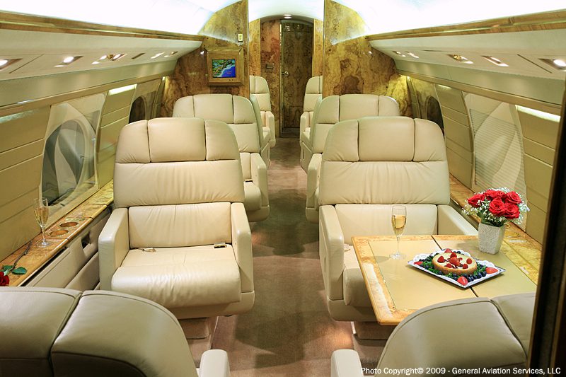 Luxurious private jet interior with beige leather seats, wood accents, a table set with flowers, fruit, and champagne glasses.