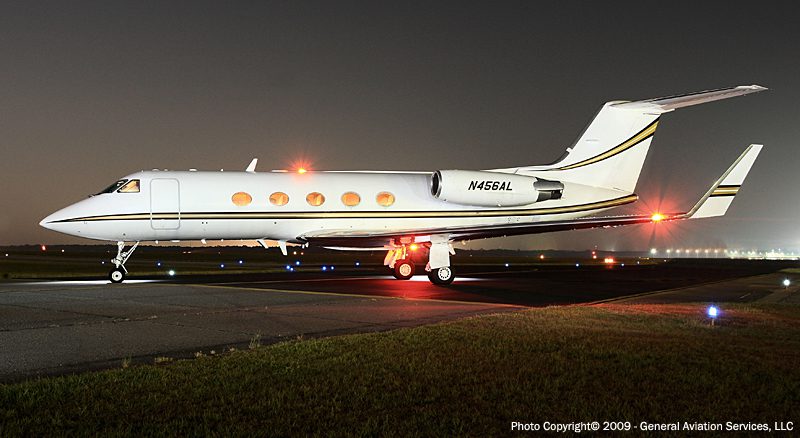 A white private jet with the registration N456AL sits on an airport runway at night, illuminated by exterior lights.