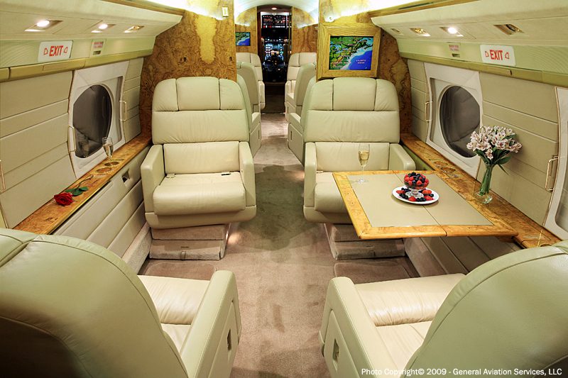 Interior of a private jet with cream leather seats, a wooden table set with a drink and dessert, and windows along the walls.