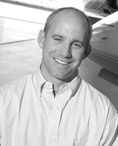A man with short hair and a button-down shirt smiles at the camera while standing in front of an aircraft.