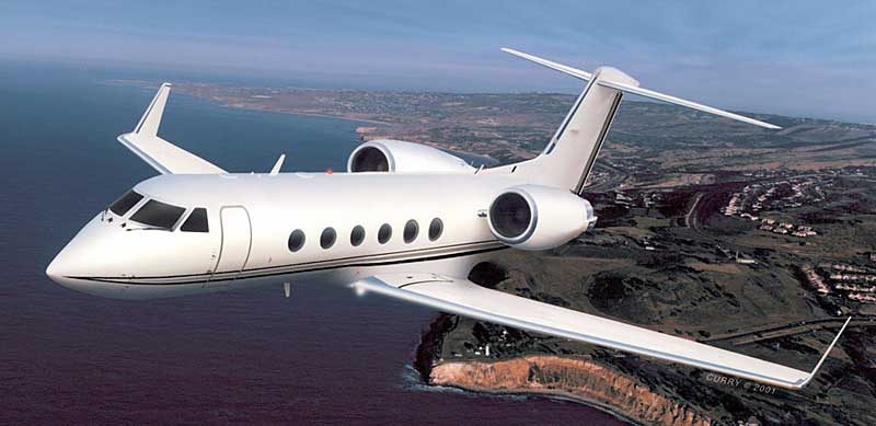 A white private jet flies above a coastal landscape with cliffs, ocean, and buildings visible below.