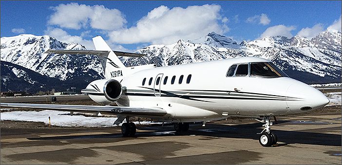 A 1991 Hawker 1000 used corporate business jet with ESP Gold Engine Program is parked on an airport tarmac, set against snow-covered mountains and a partly cloudy sky. This jet is for sale and features Domestic WiFi.