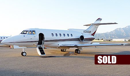 A white private jet with an open door is parked on the tarmac. A "SOLD" label is displayed in the lower right corner. Mountains are visible in the background.