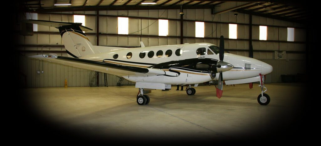 A white and black twin-turboprop aircraft is parked inside a spacious hangar with a concrete floor and metal roof.