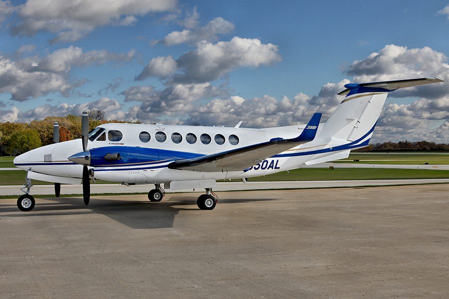 A white and blue twin-turboprop aircraft is parked on a concrete apron with grass and trees in the background under a partly cloudy sky.