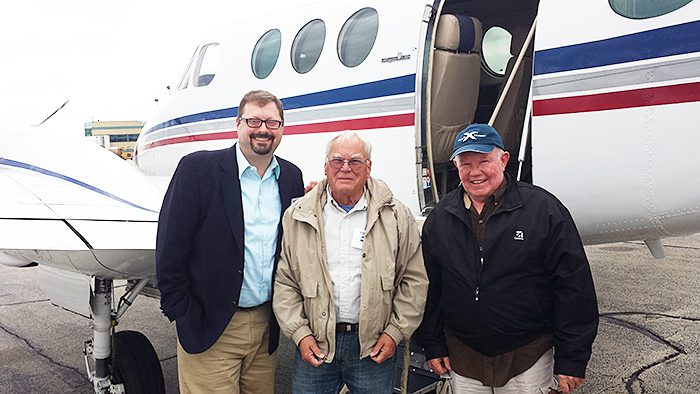 Three men stand in front of a 1979 King Air E90 H80 Conversion, just sold by General Aviation Services, smiling at the camera on an overcast day. The small white airplane features bold red and blue stripes.