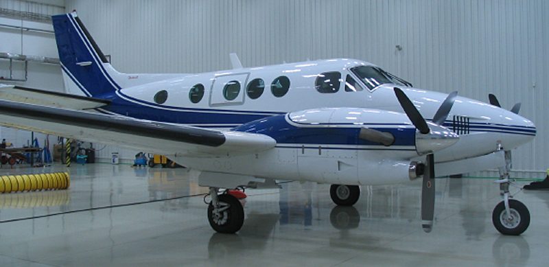 A twin-engine propeller aircraft with blue and white livery is parked inside an aircraft hangar.