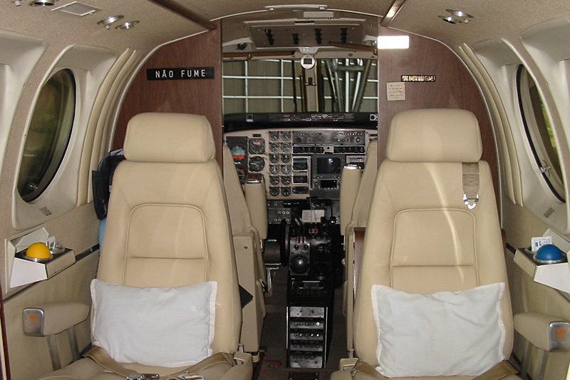 Cockpit and beige leather passenger seats inside a small private jet, with control panels visible beyond the seats and signs displayed on the wall.