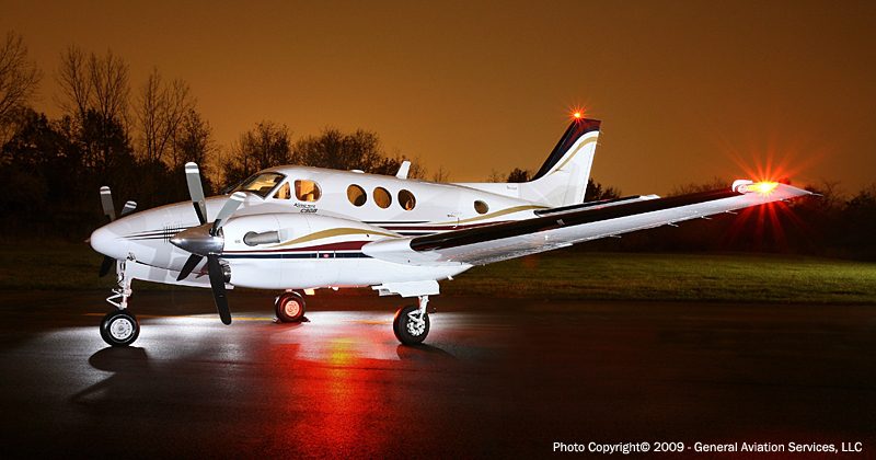 A white twin-engine propeller airplane with red and gold stripes sits on wet pavement at night, illuminated by external lights.