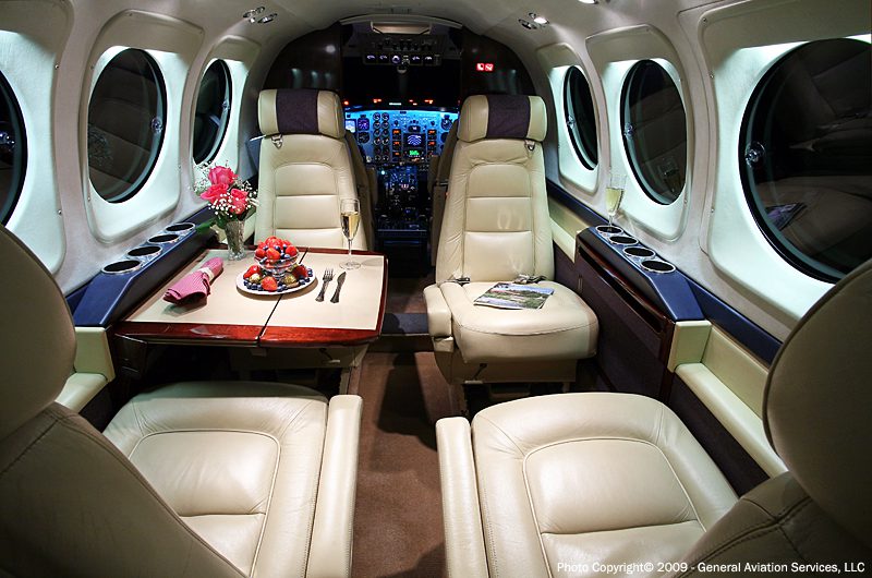 The interior of a private jet with cream leather seats, a table set with fruit, champagne, and flowers, and a cockpit visible at the front.