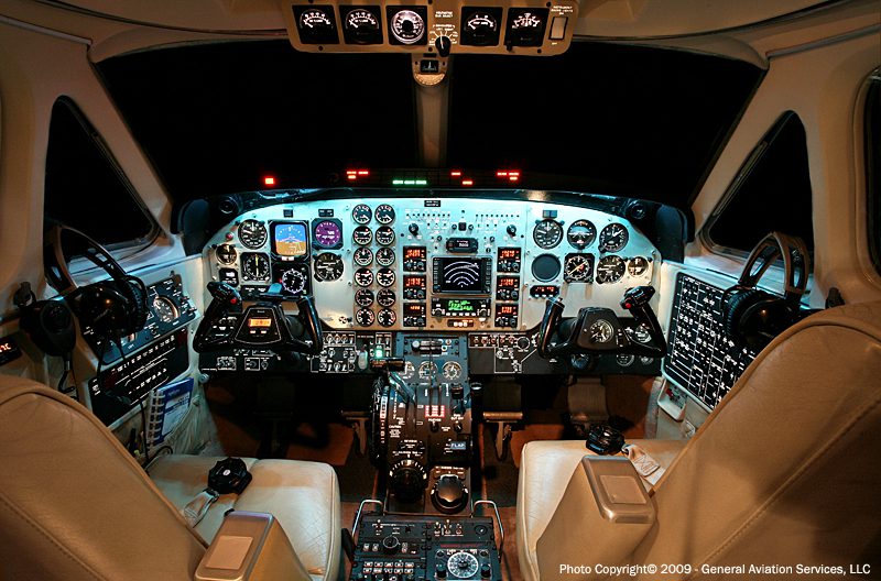 Cockpit interior of an aircraft with illuminated control panels, dual pilot seats, numerous gauges, switches, and flight instruments visible.