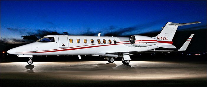 A 2001 Learjet 45, serial number 0187 and sold by General Aviation Services, with red stripes is parked on a runway at dusk, set against a striking dark blue and orange sky.