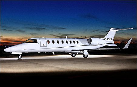 A white Learjet 45XR is parked on a runway at dusk, its lights illuminating the aircraft; the sky is dark blue with orange hues near the horizon—a stunning view for anyone seeking a used Lear.