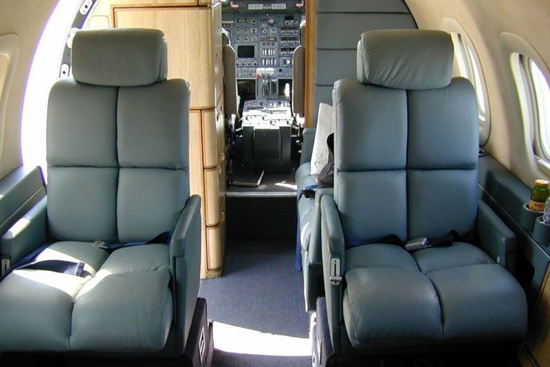 Two leather seats inside a private jet, with the cockpit and control panels visible in the background.