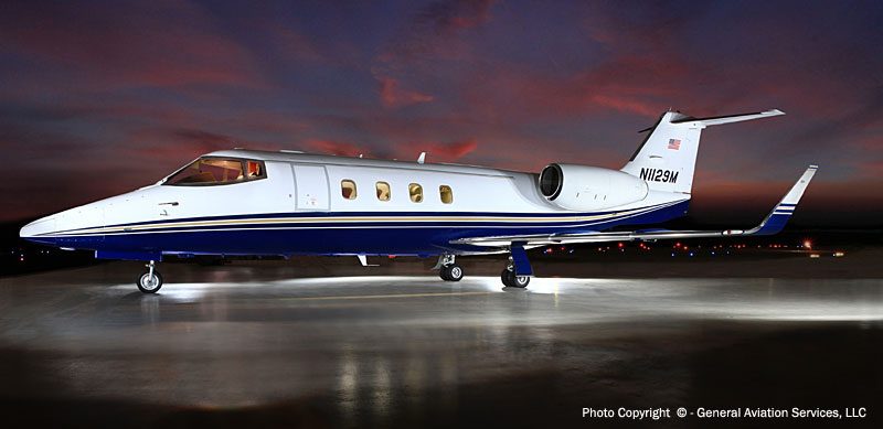 A white and blue private jet is parked on a lit tarmac at dusk, with a dark sky and faint clouds in the background.
