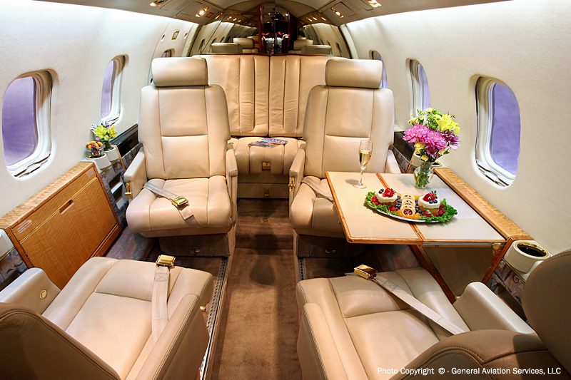 The image shows the luxurious interior of a private jet with beige leather seats, a fold-out table set with snacks, flowers, and drink, and windows along the cabin walls.