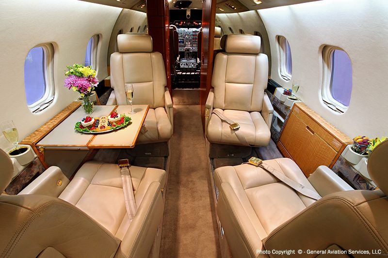 Interior of a private jet with beige leather seats, a table set with food, flowers, and drinks, and a cockpit visible at the front.