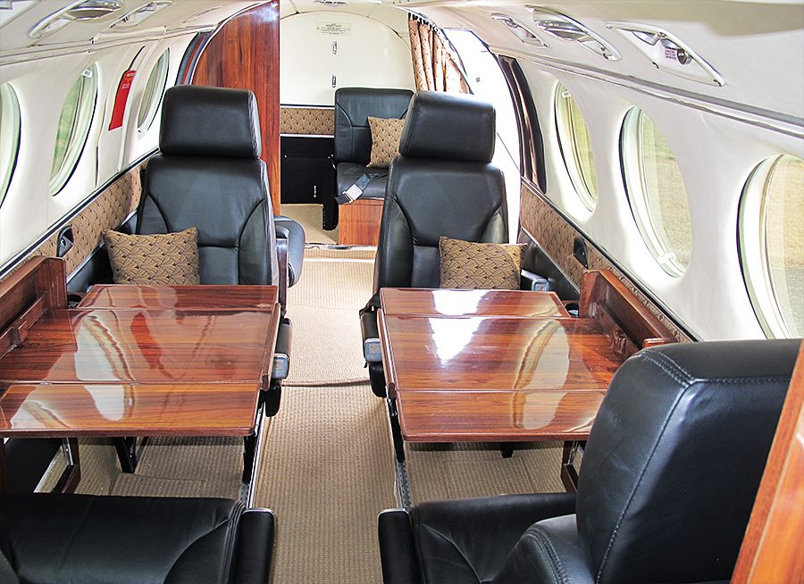 Interior of a private jet with four leather seats and two wooden tables, side windows, beige carpet, and decorative pillows.