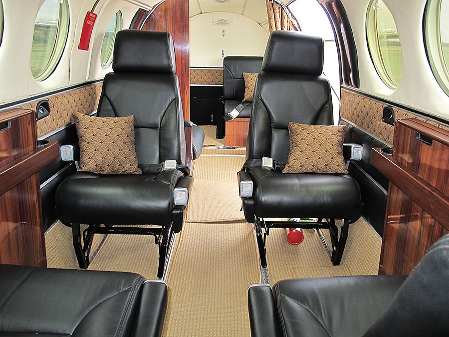 Interior of a private jet showing four black leather seats, patterned pillows, wood paneling, and round windows.