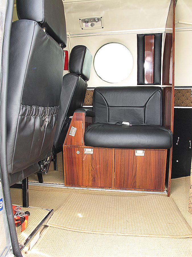 The image shows the interior of a small aircraft, featuring leather seats, wood-paneled storage, and beige carpet flooring. A round window is visible on the side wall.