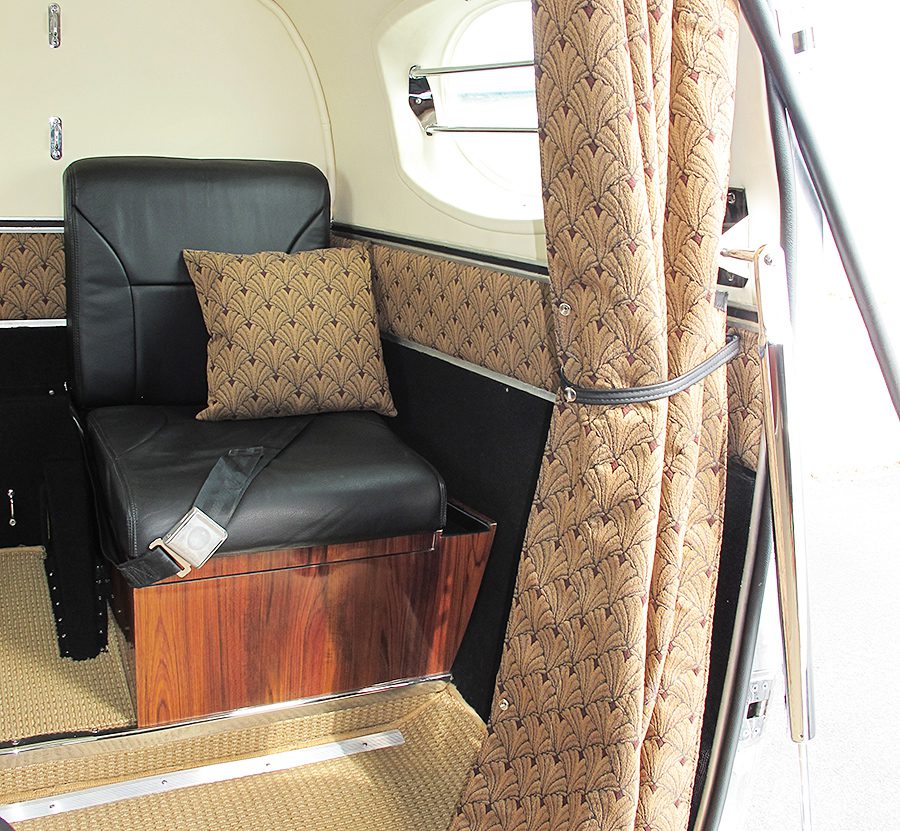 A single black leather seat with a matching patterned cushion inside a small, vintage cabin with patterned curtains and beige carpeting.