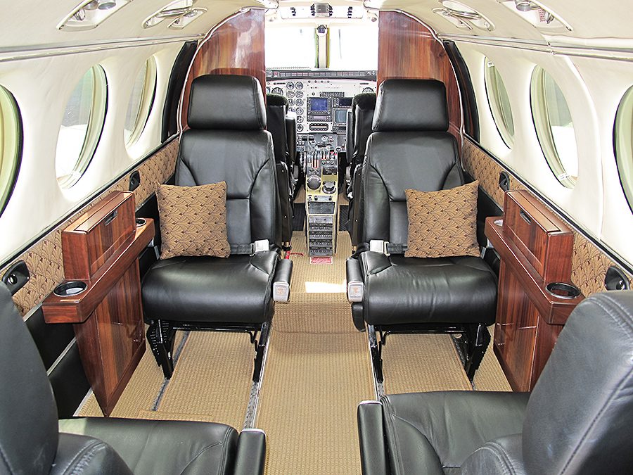 Interior of a small private jet with four black leather seats, wooden side panels, beige carpet, and cockpit controls visible at the front.
