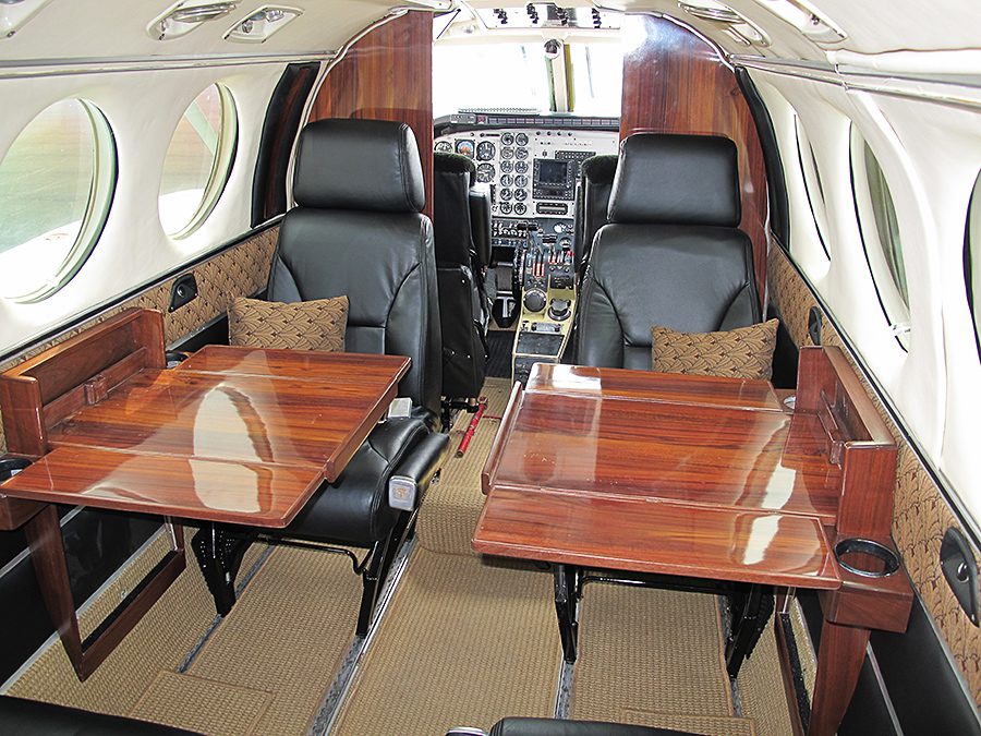 Interior of a private jet featuring leather seats, fold-out wooden tables, and cockpit controls visible at the front.