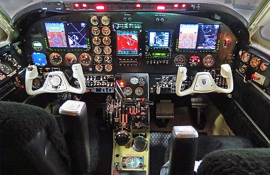 Cockpit view of an aircraft showing dual control yokes, multiple digital displays, analog gauges, switches, and illuminated indicator lights on the instrument panel.