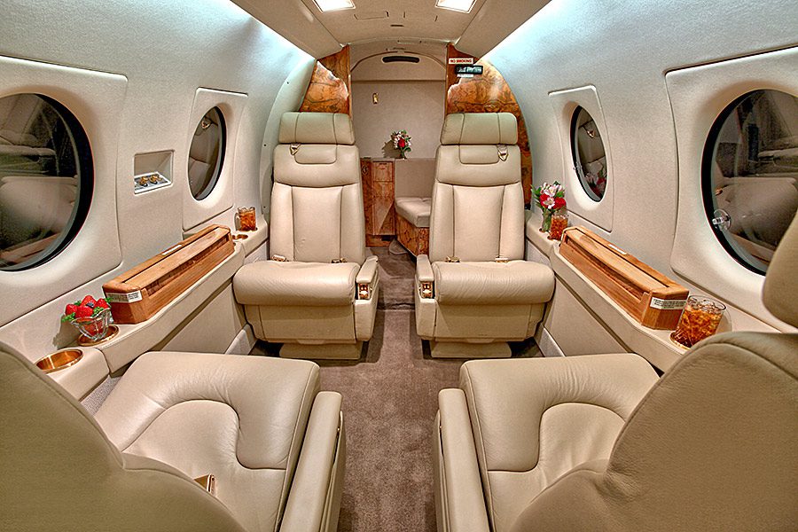 Interior of a private jet with four cream leather seats, wood paneling, round windows, and small tables with drinks and flower arrangements.