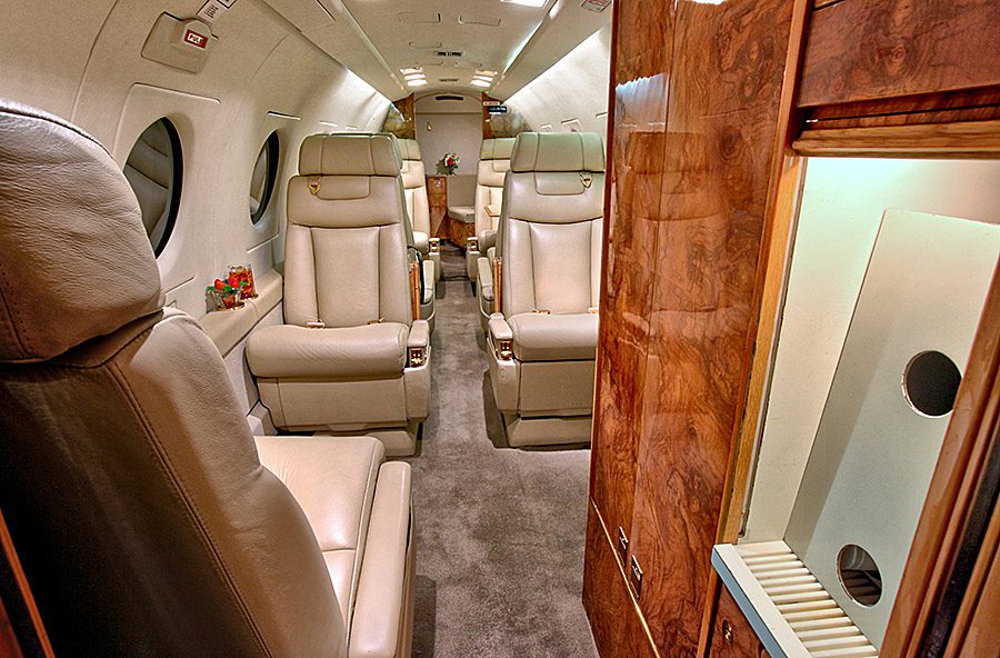 Interior of a private jet with four cream leather seats, wood paneling, carpeted floor, side windows, and a small vase with flowers on a side ledge.