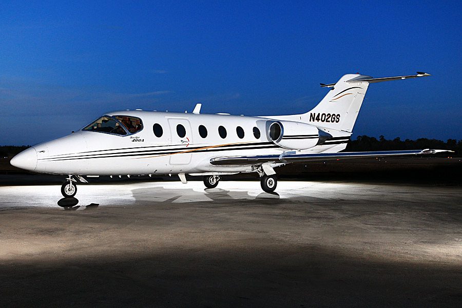 A white private jet with tail number N402QS is parked on a lit tarmac at dusk, with a dark blue sky in the background.