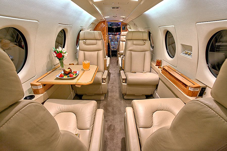 Interior of a private jet with cream leather seats, a table set with food and flowers, and a view of the cockpit in the background.