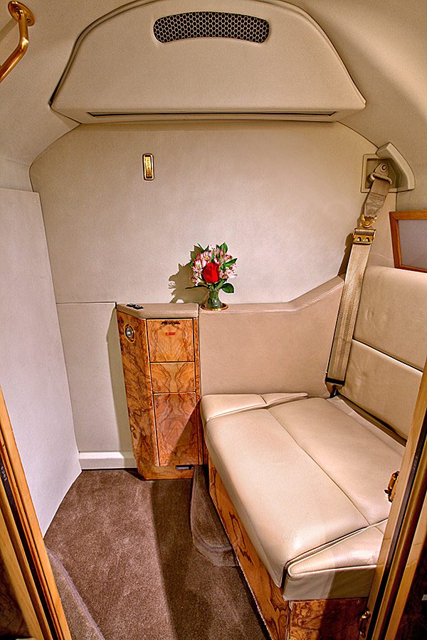 A small, private jet cabin with a beige leather seat, wooden side cabinet, and a vase of flowers on top of the cabinet.