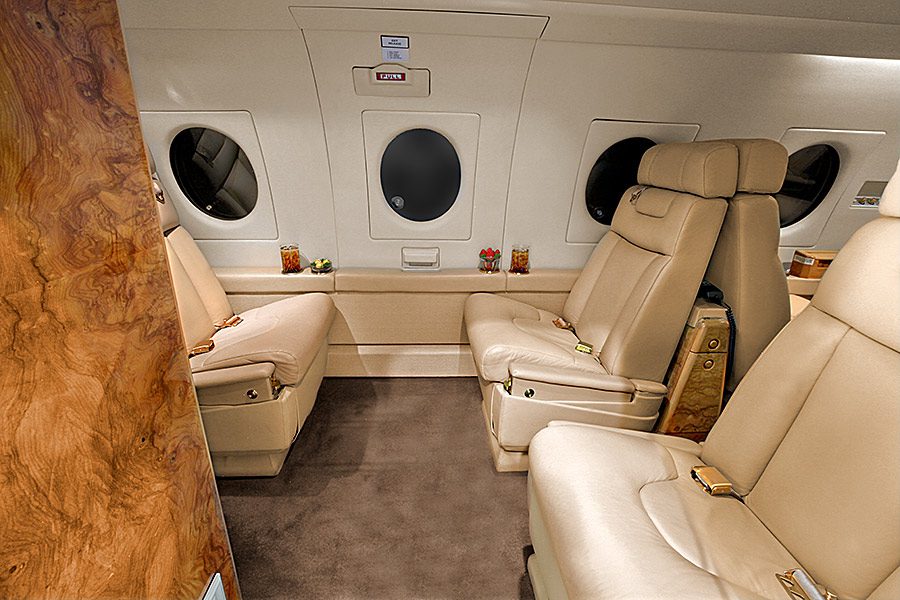 Four beige leather seats facing each other inside a private jet cabin, with drinks on side tables and oval windows on the wall.