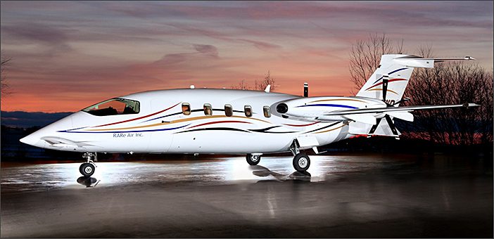 A white Used Piaggio P180 Avanti business aircraft with red, gold, and blue stripes is parked on a wet tarmac at sunset, with bare trees in the background—recent price reduction to $2,195.