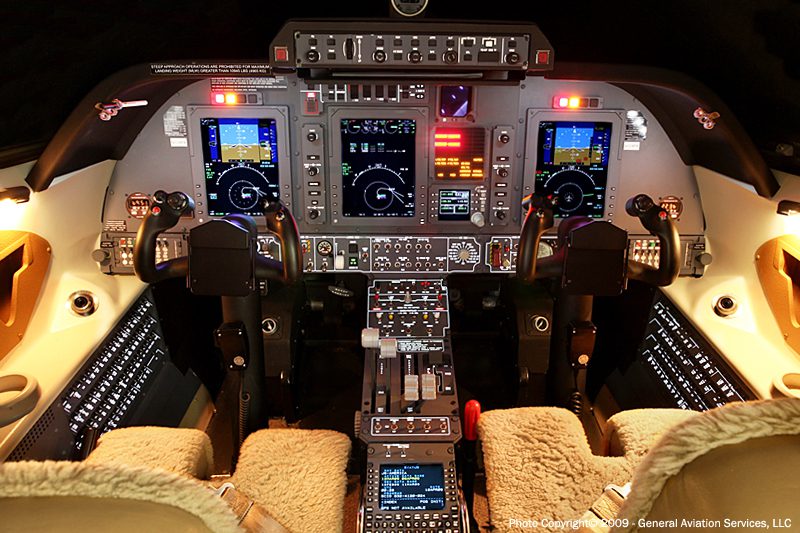 Cockpit view of a modern aircraft with illuminated control panels, dual flight displays, numerous switches, and two pilot seats with sheepskin covers.