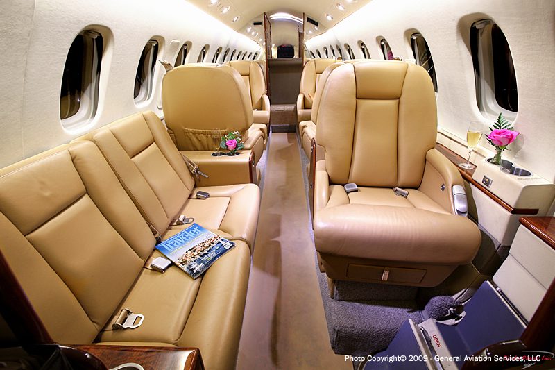 Interior of a private jet with tan leather seats, small tables with pink flowers, magazines, and overhead lighting.
