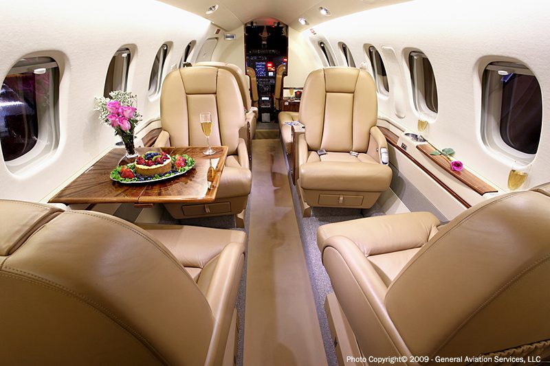 The interior of a private jet with four beige leather seats, a table with a meal and flowers, and glasses of champagne near the windows.