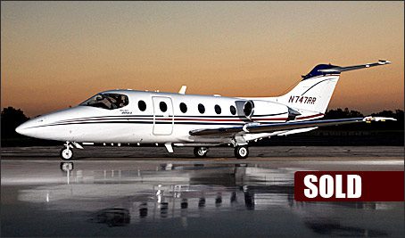 A small white private jet with blue and red stripes is parked on a runway at dusk. A "SOLD" label appears in the lower right corner.