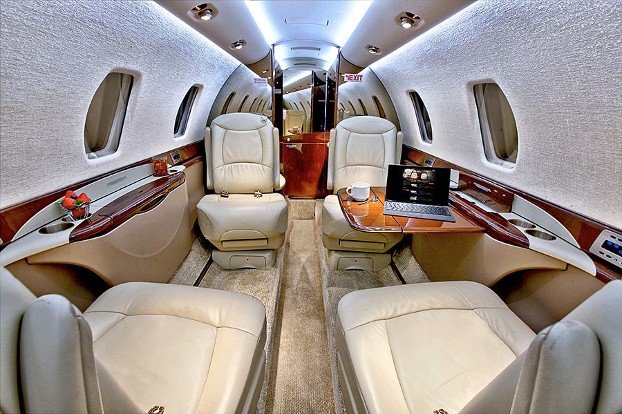 Interior of a private jet featuring four cream leather seats, wooden accents, side tables, and a laptop on a tray.