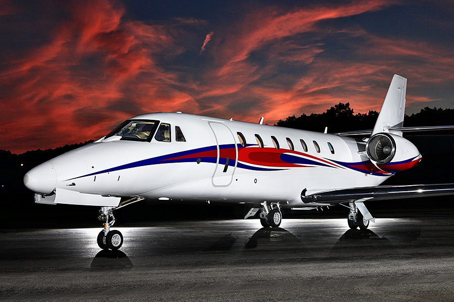 A white private jet with blue and red stripes is parked on a tarmac at dusk, with a dramatic red and purple sunset sky in the background.