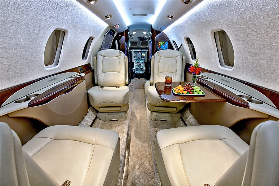 The interior of a private jet with four cream leather seats, a fold-out table holding a drink and fruit, and a view toward the cockpit.
