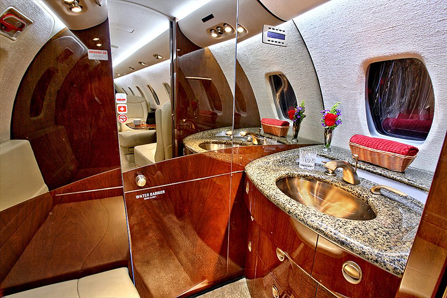 Private jet bathroom with a granite countertop, stainless steel sink, mirror, fresh flowers, and a wicker basket, featuring wooden cabinetry and an oval window.