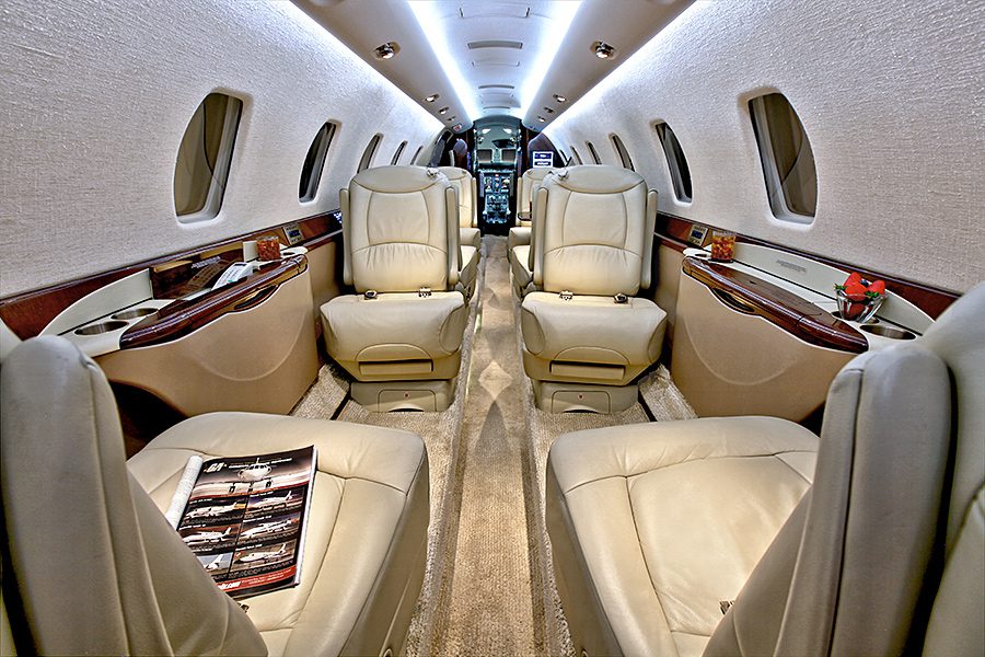 Interior of a private jet showing cream leather seats, wood paneling, a magazine on a seat, and the cockpit visible at the far end of the aisle.