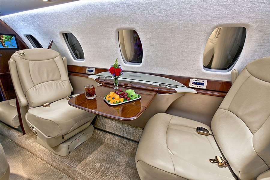 Two cream leather seats in a private jet cabin with a table set with fruit, a drink, and a vase with flowers between them.