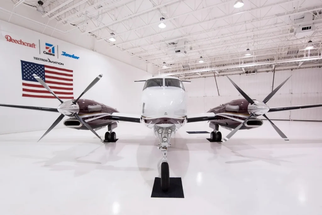 A twin-propeller airplane is parked inside a brightly lit hangar, with an American flag and aviation company logos visible on the wall behind it.