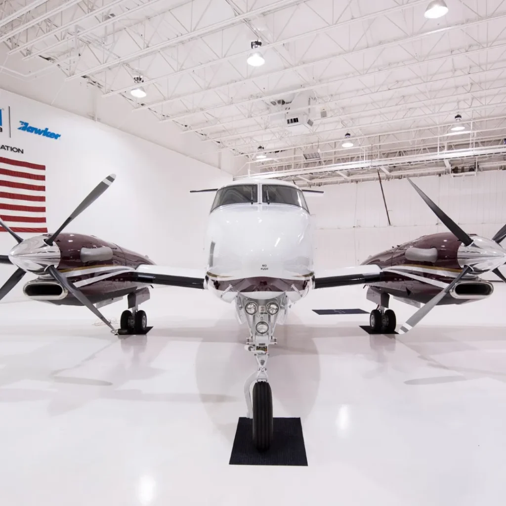 A twin-engine propeller aircraft is parked inside a well-lit hangar with an American flag on the wall in the background.