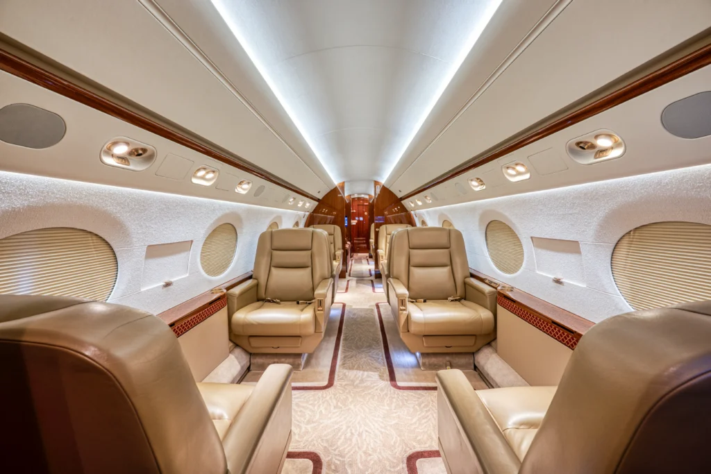 Interior view of a private jet showing four beige leather seats, carpeted floor, wood accents, and soft overhead lighting.