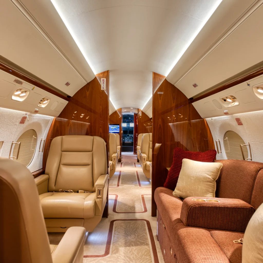 Interior of a luxurious private jet featuring tan leather seats, plush carpeting, polished wood paneling, and a view of the cockpit at the far end.