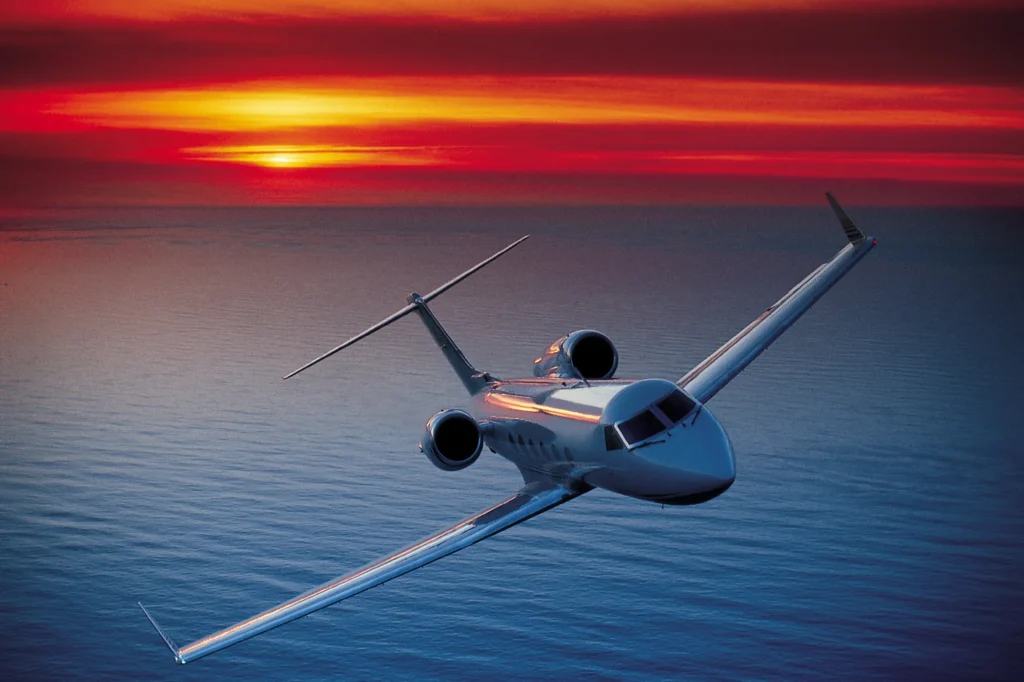 A private jet flies over the ocean at sunset, with an orange and red sky reflecting on the water.